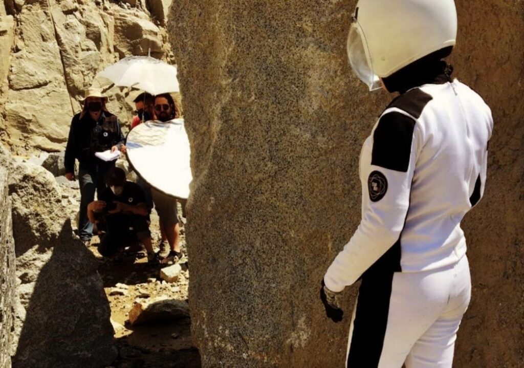 Imagem de bastidores mostrando uma equipe de filmagem trabalhando em uma cena com um ator vestido de astronauta em um ambiente rochoso. A equipe usa equipamentos como refletores e guarda-sol para controlar a iluminação, enquanto o ator, de costas para a câmera, observa a cena.