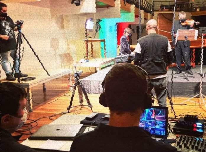 Cena dos bastidores de uma gravação, com uma equipe técnica ajustando câmeras, equipamentos de som e iluminação. Pessoas com fones de ouvido monitoram a gravação em telas, enquanto um músico toca violino ao fundo. A configuração sugere uma produção audiovisual profissional em ambiente interno.