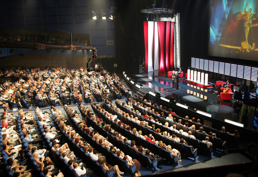 Auditório lotado durante evento de cinema, com público sentado em fileiras observando uma tela gigante projetada na frente do palco